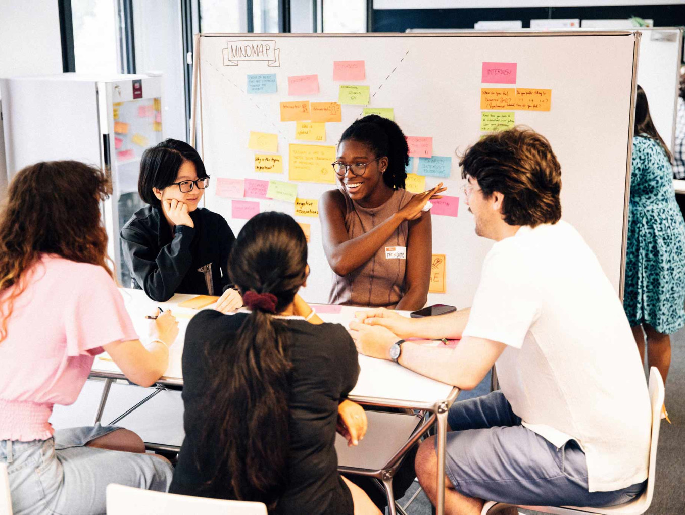 A group sitting in a circle while talking and laughing in front of a wall with Post-its on it.