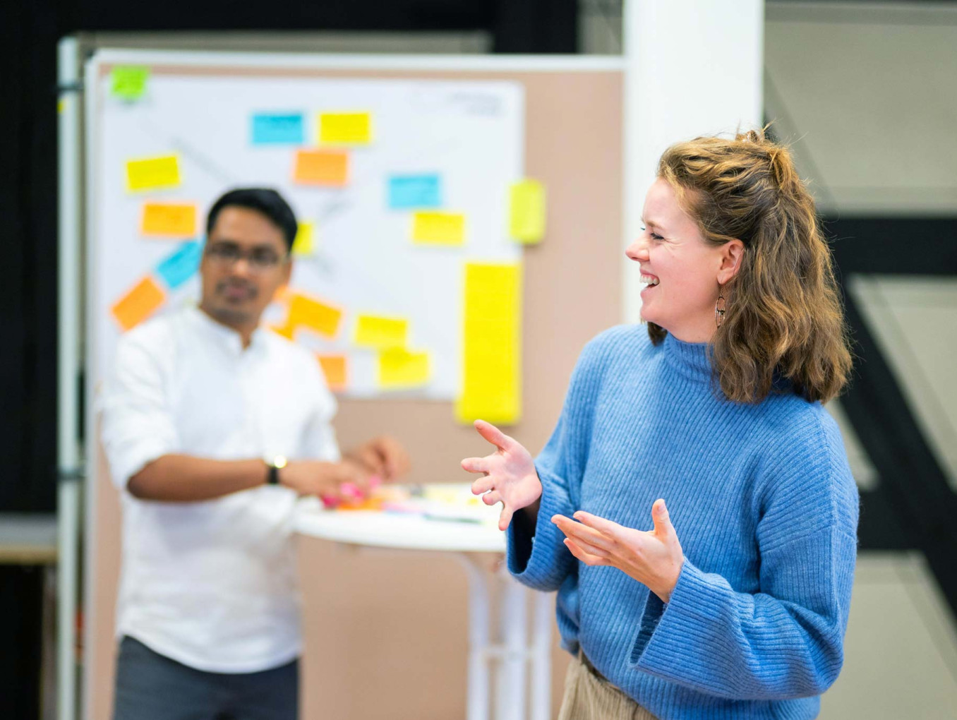 A person speaks and gestures during a workshop discussion, while another participant stands near a board covered with colorful sticky notes.