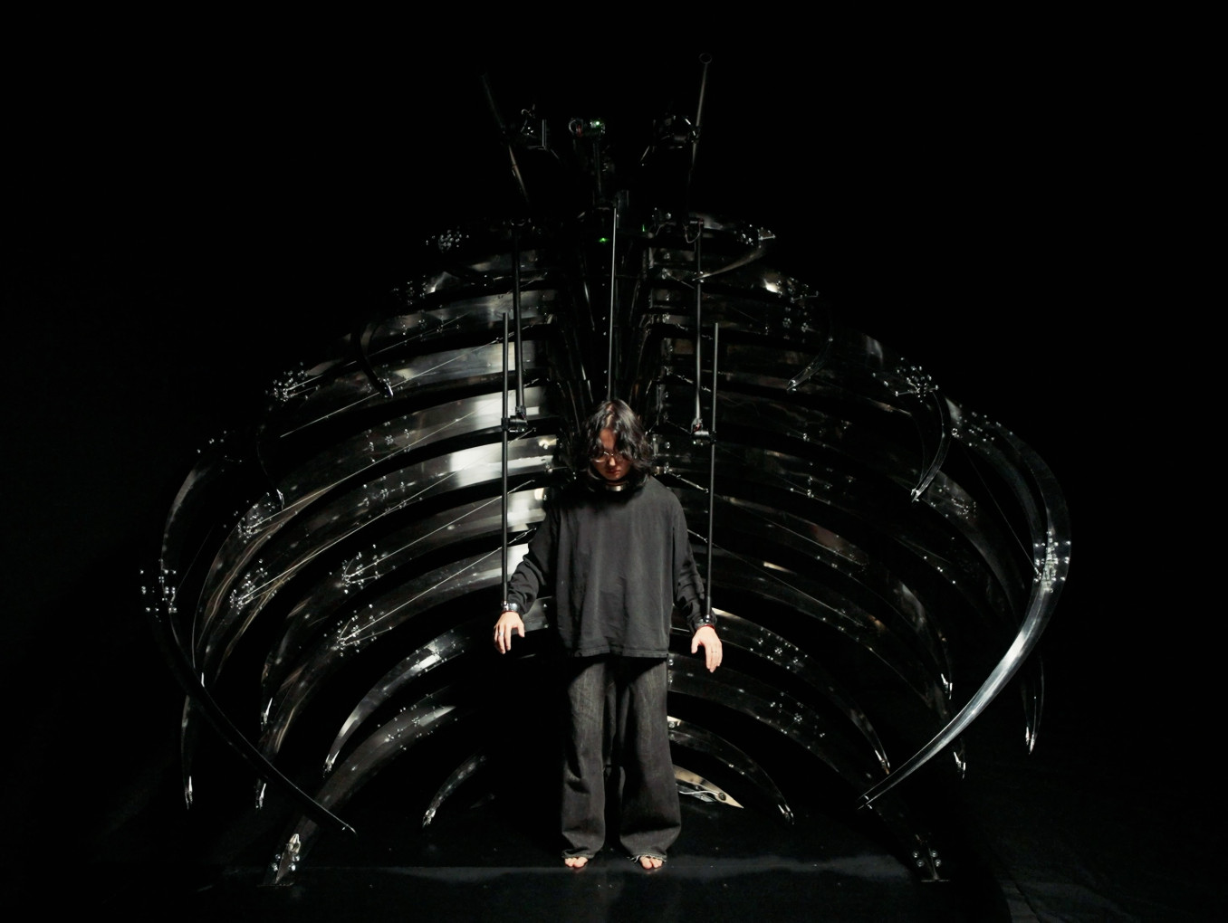 A participant stands inside xoxo-skeleton, a polished steel ribcage 2.5 metres tall and 3 metres in diameter. Twenty-four independent language model agents read the surroundings through 480 sensor channels, deliberate among themselves through MaryGPT, a language model fine-tuned on the complete text of Mary Shelley's Frankenstein, and move the participant's wrists and neck through three ceiling-mounted robotic arms.