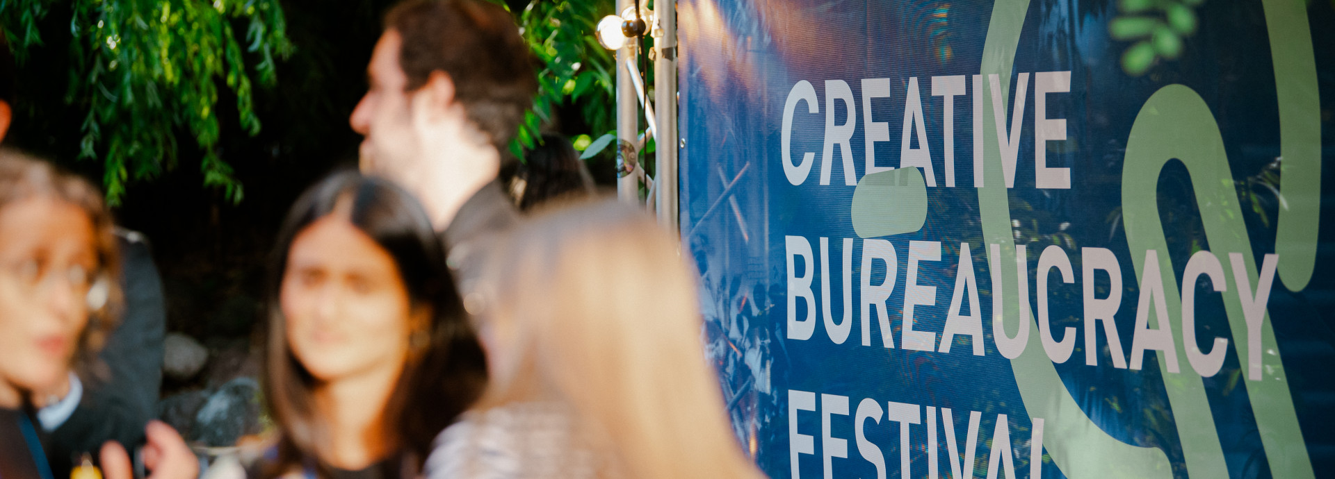people at the festival area outside standing and talking infront of a big poster of the festival 