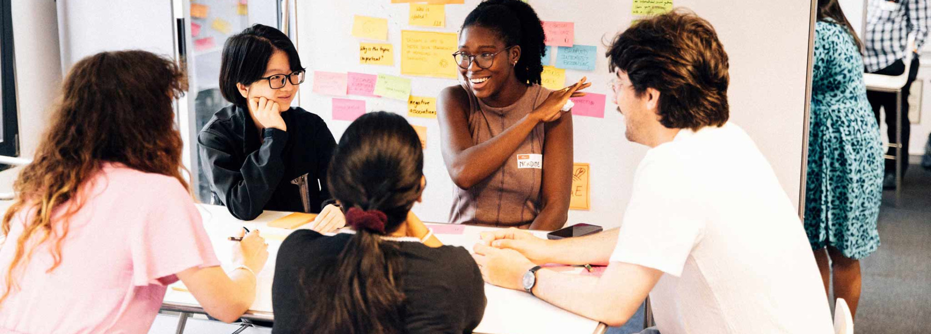 A group sitting in a circle while talking and laughing in front of a wall with Post-its on it.