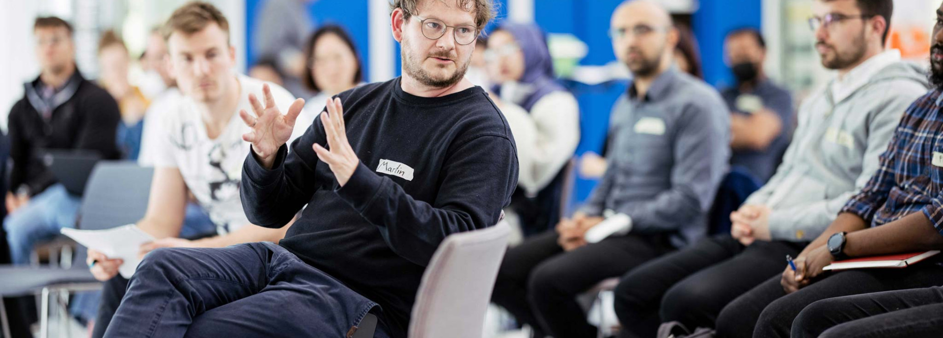 A person speaks while gesturing with hands during a group discussion in a workshop setting.