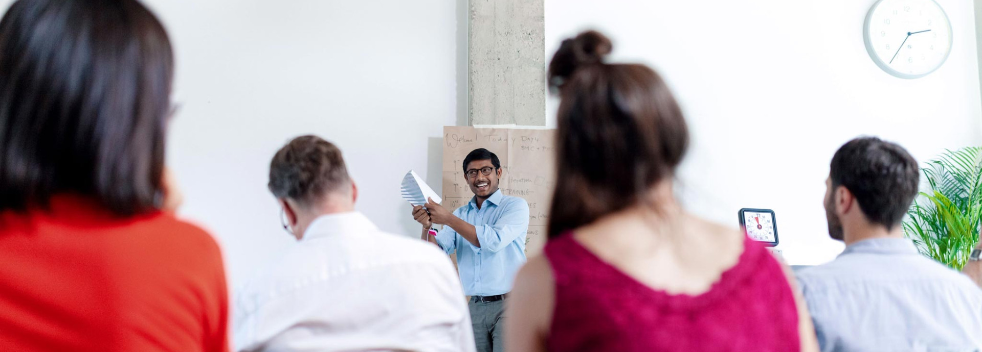 A person is standing in front of a group of seated individuals, holding papers and speaking with a smile.