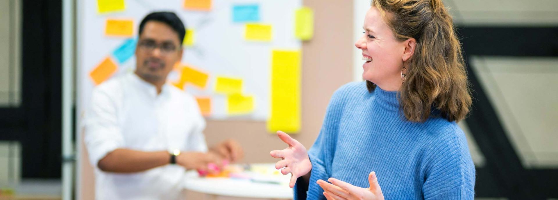 A person speaks and gestures during a workshop discussion, while another participant stands near a board covered with colorful sticky notes.