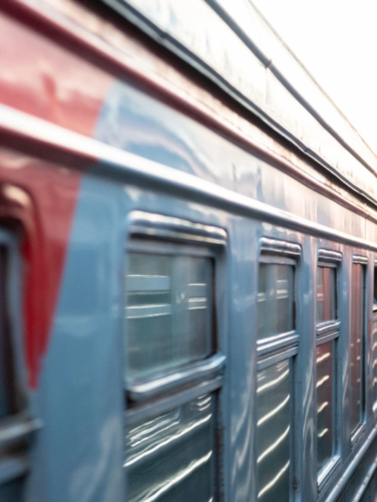 A side view of two trains, one on the left in focus, appearing to be stationary or moving slowly parallel to each other. The train on the left has red and light blue stripes along its side with reflective silver details around its windows. The train on the right is mostly obscured, showing only a narrow side profile, creating a tight space between the two. The perspective is angled, with the trains stretching into the distance under a bright, possibly overcast, sky.