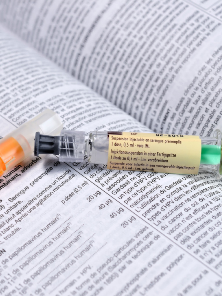 A close-up shot of two pre-filled syringes resting on an open medical or pharmaceutical document filled with text. The syringe on the left has an orange plunger and cap, while the syringe on the right has a green plunger, a yellow label with text, and a clear barrel containing liquid. The text on the document is in multiple languages, likely detailing drug information or instructions.