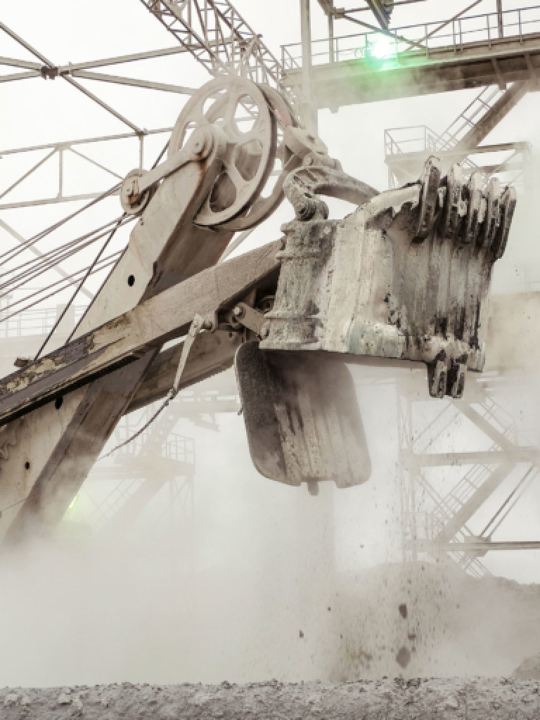 A large industrial machine, possibly a bucket-wheel excavator or similar heavy machinery, with a massive bucket attachment, is actively working amidst a cloud of dust or steam. The machine is grey and appears worn, suggesting a harsh working environment. In the background, other parts of the industrial structure and sky are visible, obscured by the haze.