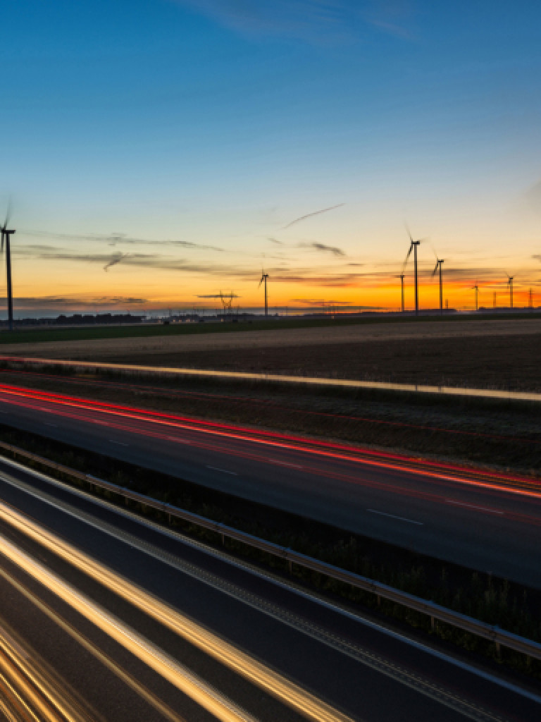 A long exposure photograph captures a highway at dusk with streaks of light from vehicle headlights and taillights. On the right, a field is dotted with numerous wind turbines, some with blurred blades indicating motion. Power lines are visible in the distance on the left. The sky transitions from a deep blue at the top to warm oranges and yellows near the horizon, illuminated by the setting sun.