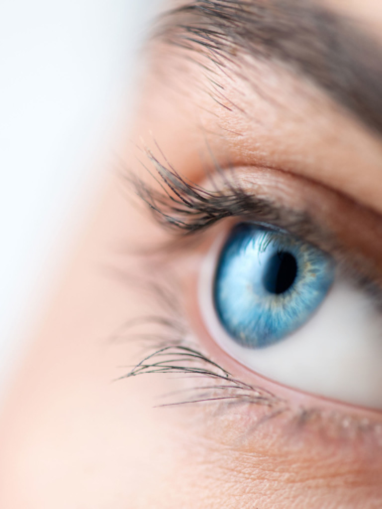 A close-up, side-profile shot of a human eye with striking blue irises, looking upwards and slightly to the right. Eyelashes are visible, and the eyebrow is partially in frame at the top. The skin around the eye is detailed, and the background is a soft, bright blur, putting the focus entirely on the eye.