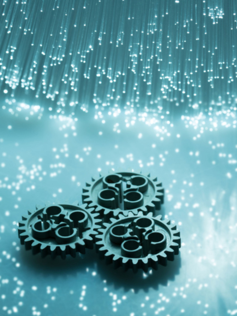 A visually striking image featuring three dark, metallic gears positioned on a light blue, shimmering surface. Above them, a cluster of thin, illuminated fiber optic strands descends, creating a sparkling, ethereal effect. Small, bright dots are scattered across the surface, enhancing the luminous and technological theme. The overall impression is one of connection, technology, and precision.