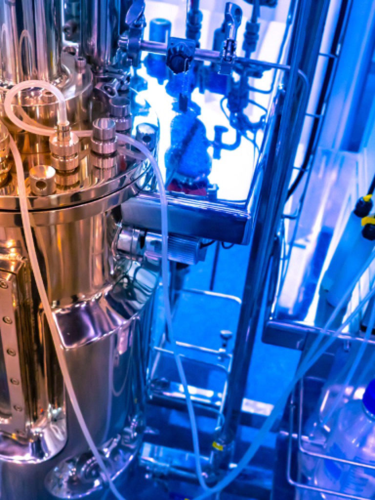 A high-angle close-up shot of a large, gleaming stainless steel bioreactor or fermentation vessel, indicating a scientific or industrial biotech setting. Numerous pipes, tubes, gauges, and valves are connected to the main vessel, with clear tubing carrying liquids. The background features more scientific equipment and machinery, bathed in cool blue and white light, reflecting off the metallic surfaces. A small clear bottle is visible in a tray at the bottom right.