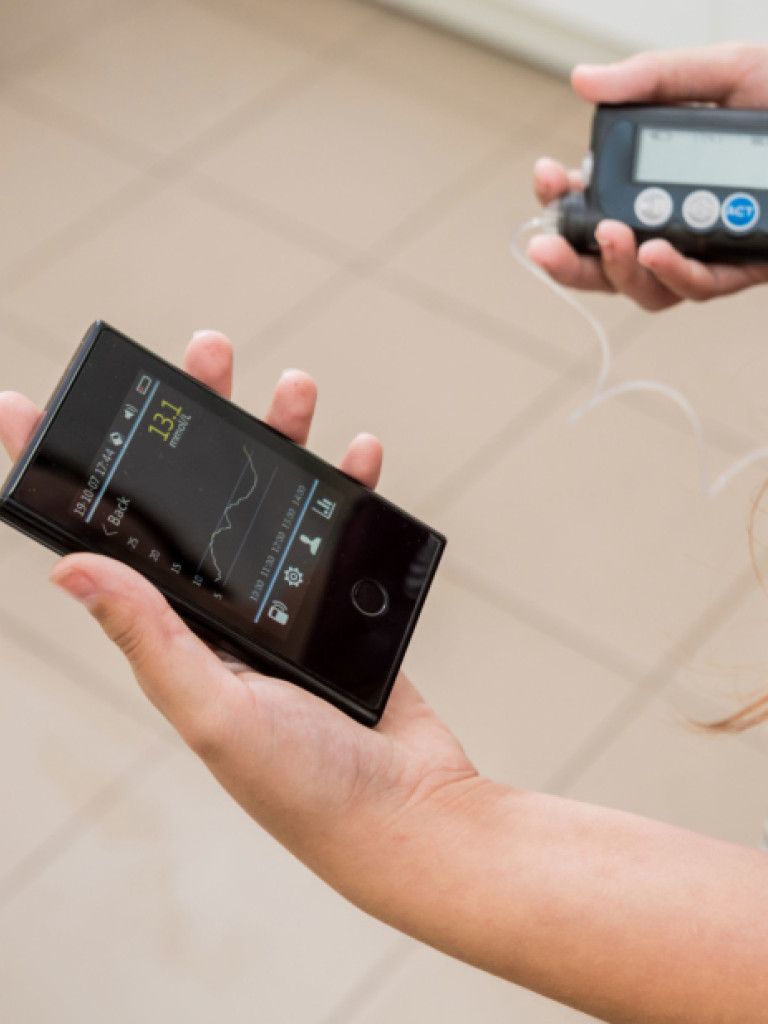 A person is holding two medical devices, likely for diabetes management. In the left hand, there's a sleek, black smartphone-like device displaying a numerical reading of "13.1 mmol/L" and a graph, along with icons. In the right hand, a darker, more rugged device with buttons and a small screen is held, possibly an insulin pump or a continuous glucose monitor transmitter, with tubing extending from it. The background is a blurred, light-colored tiled floor.