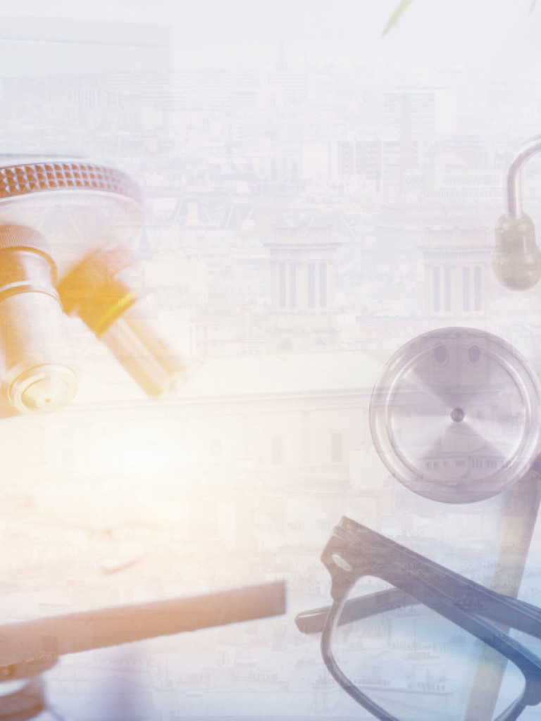 A soft, light-filled, double-exposure image combines elements of medical science and urban architecture. In the foreground, a close-up of a microscope's objective lenses is visible on the left, bathed in a warm glow. On the right, medical items like a stethoscope, a pen, and a pair of glasses are arranged next to a clipboard with a "MEDICAL" heading and an "Rx" symbol. Superimposed over these elements, and more distinctly in the background, is a faded, monochromatic image of a city skyline with buildings, c