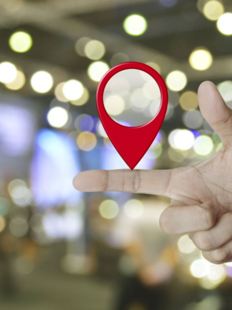A right hand is extended with the index finger pointing straight out, and a bright red, three-dimensional location pin icon is balanced on top of the finger. The background is a blurred indoor scene with numerous bokeh lights, suggesting an urban or commercial environment.