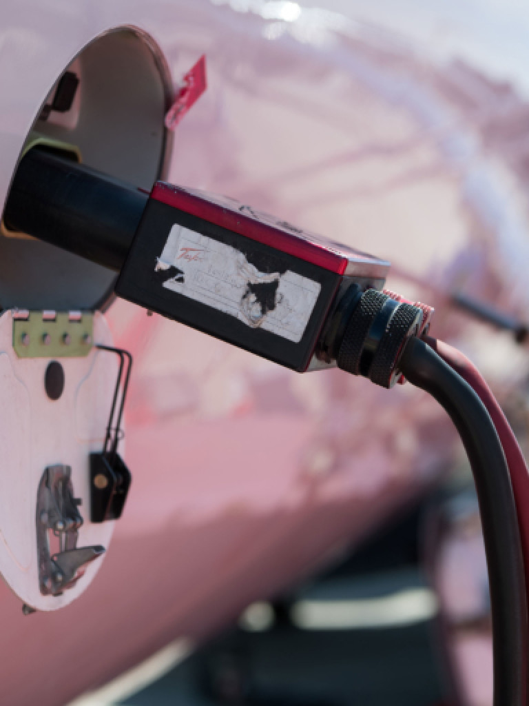 A close-up shot of a power cable being plugged into the side of a light pink or rose-colored aircraft, possibly an electric plane or eVTOL (electric vertical takeoff and landing) jet. The charging port is open, revealing internal components and some red labels. The cable itself is black with a red stripe, connecting to a rectangular black and red connector that is inserted into the port. The background is blurred, showing hints of the aircraft's structure and possibly an outdoor setting.