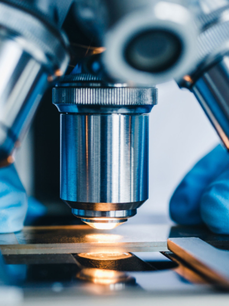 A close-up, eye-level shot focuses on the objective lenses of a microscope, which are made of shiny metal. A sample slide is visible on the stage below, illuminated by a warm, golden light coming from beneath, suggesting active observation. Two hands wearing bright blue laboratory gloves are partially visible on either side of the microscope, indicating a person is operating the equipment. The background is blurred, maintaining focus on the scientific instrument.