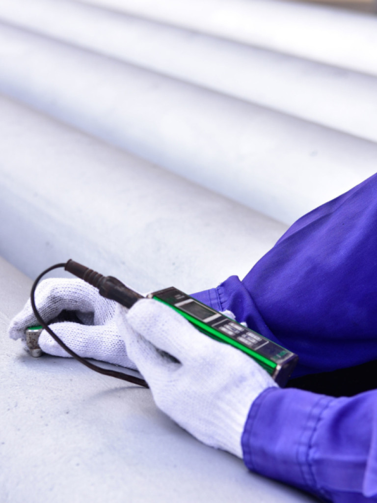gloved hands holding a measuring device. The person holding it is dressed in purplish blue work wear with a reflector stripe on the arm. The background is made of gray pipes stretching from the bottom right to the top left of the image.