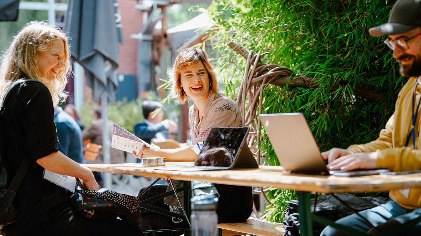 3 people sitting on a table, laughing looking at a laptop 